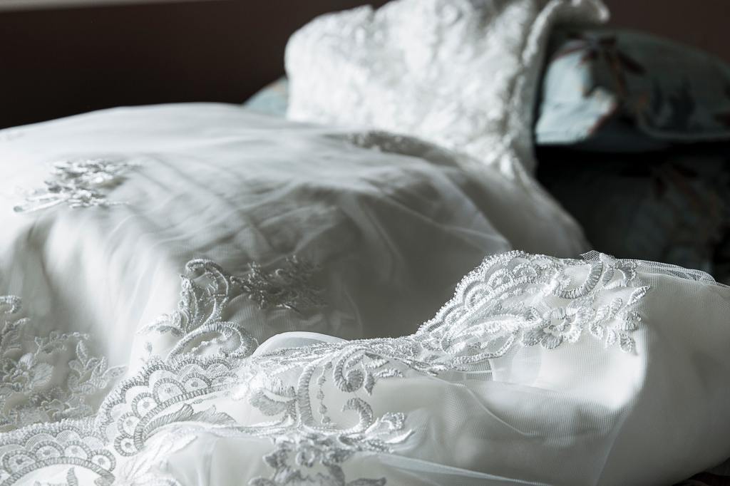 A white wedding dress laid out on a chaise lounge, the camera is focused on the  delicate embroidered lace that adorns the hem and train.