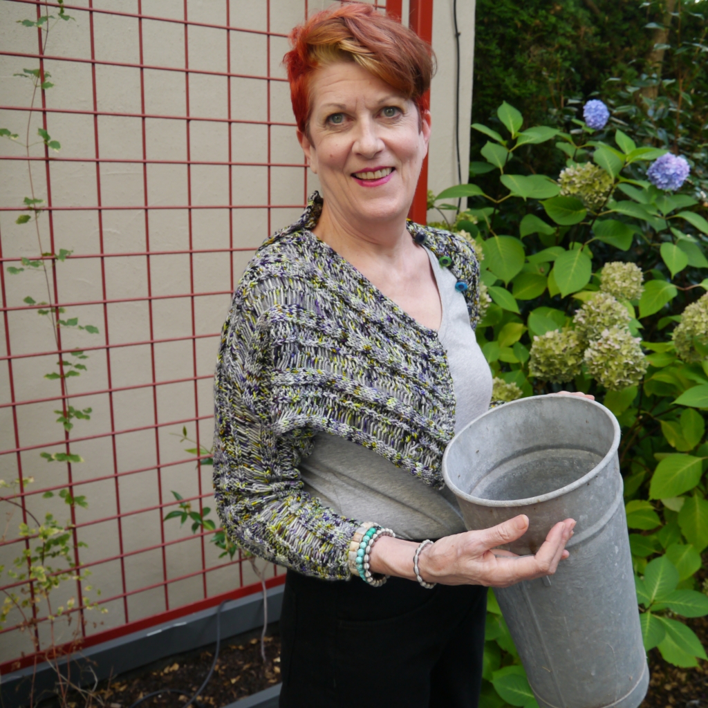 A red-headed woman wears a purple and grey shrug, with pops of yellow/green. She stands in front of a similarly colored hydrangea bush holding a french flower bucket.
