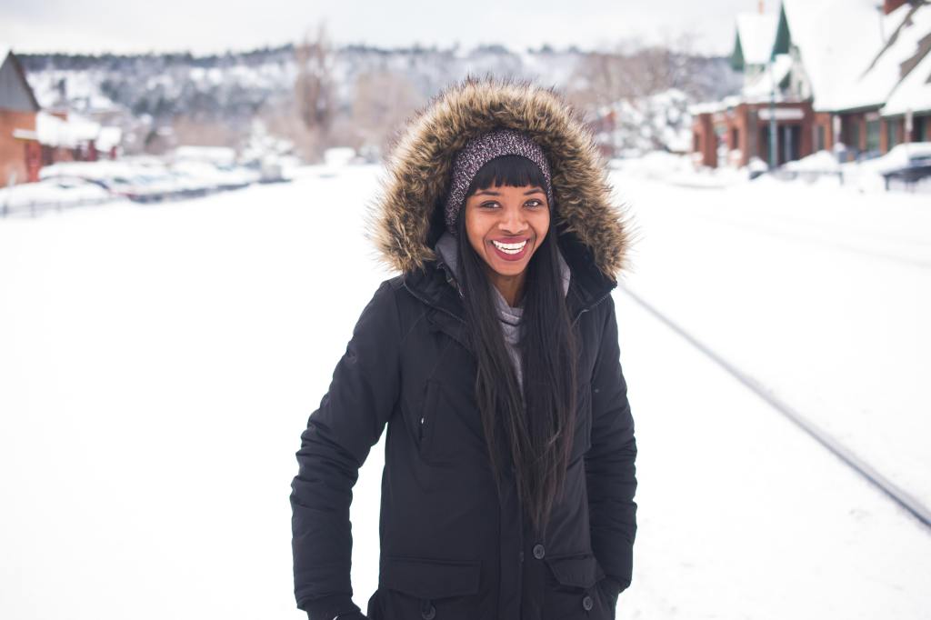 A Black woman with long black hair poses facing the camera, smiling, wearing a black coat with a faux fur hood. Behind her is a snow neighborhood.